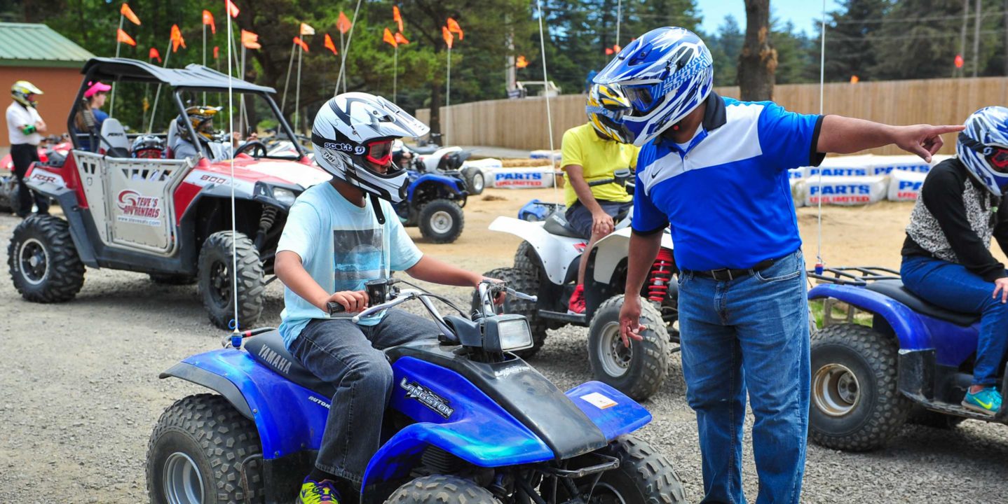 Adult talking to a child on an ATV, both wearing helmets.